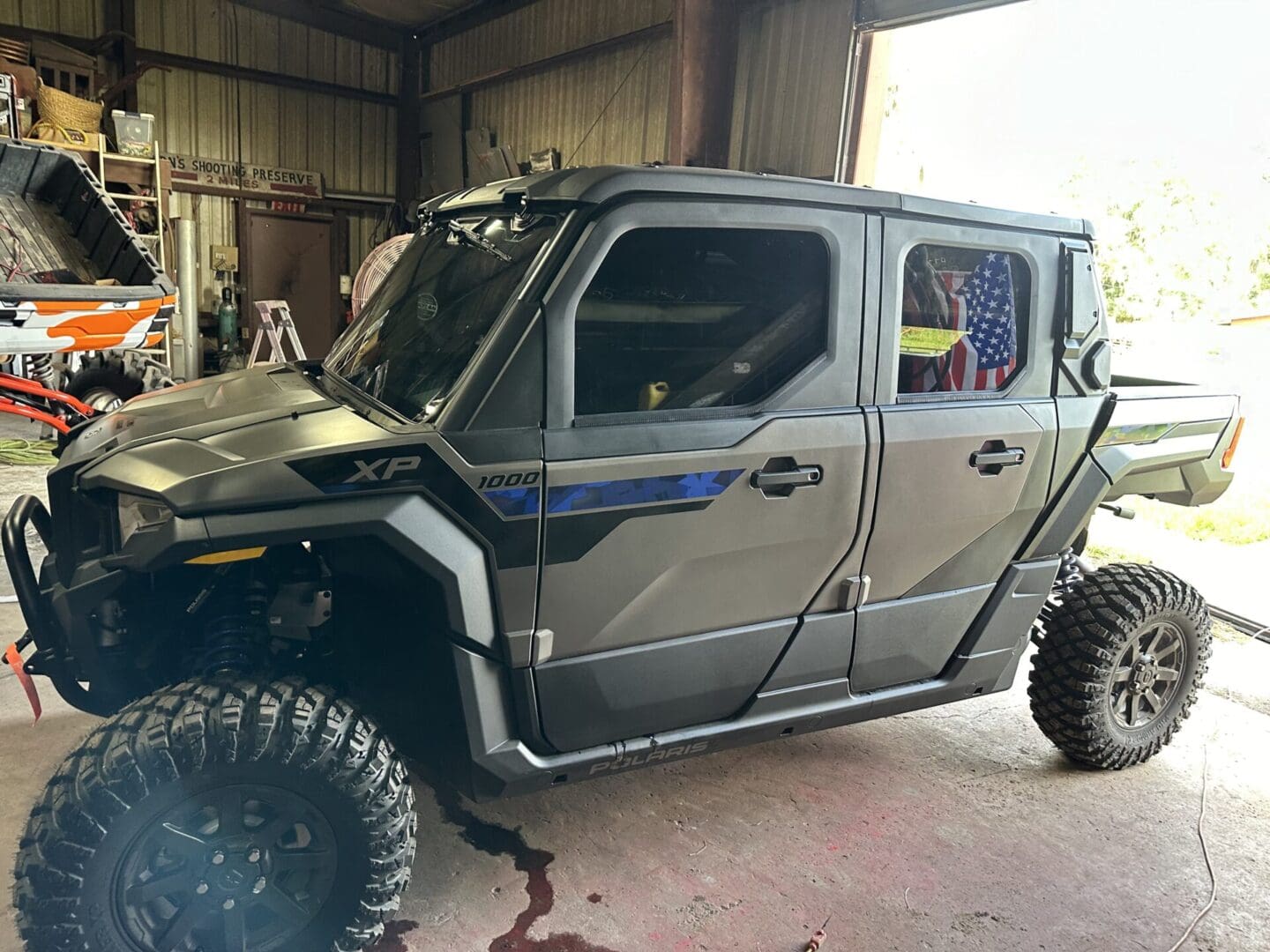 A black, rugged off-road utility vehicle is parked inside a garage with an American flag displayed in the rear window. The vehicle features large tires and a sturdy build designed for rough terrain, complemented by professional vehicle window tinting for added privacy and protection.