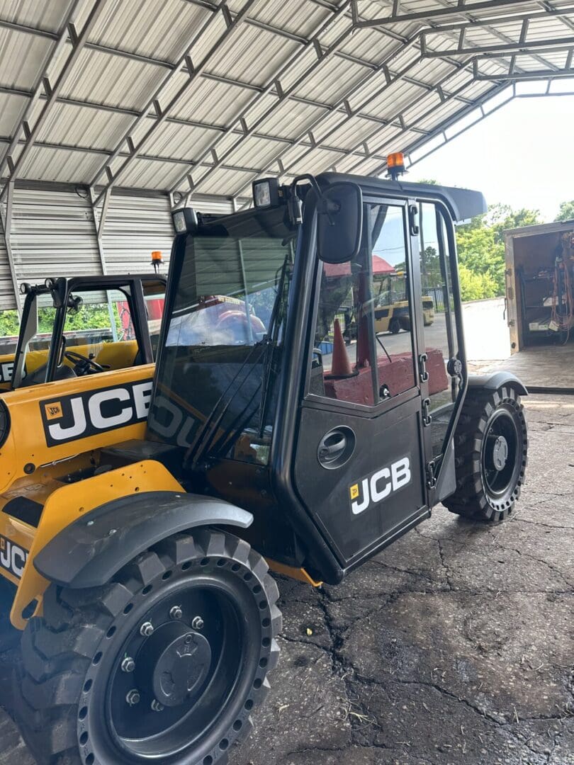 A yellow and black JCB construction vehicle, featuring vehicle window tinting, is parked under a metal canopy. The vehicle has large tires and a covered cab. Another JCB vehicle is partially visible beside it.